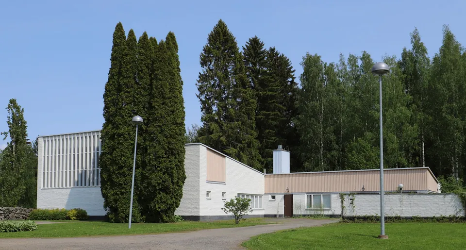 Luhangan valkotiilinen talvikirkko ja sen matala siipiosa kesäisessä pihapiirissä. Kirkkosalin ikkunoita reunustavat pystysäleiköt.